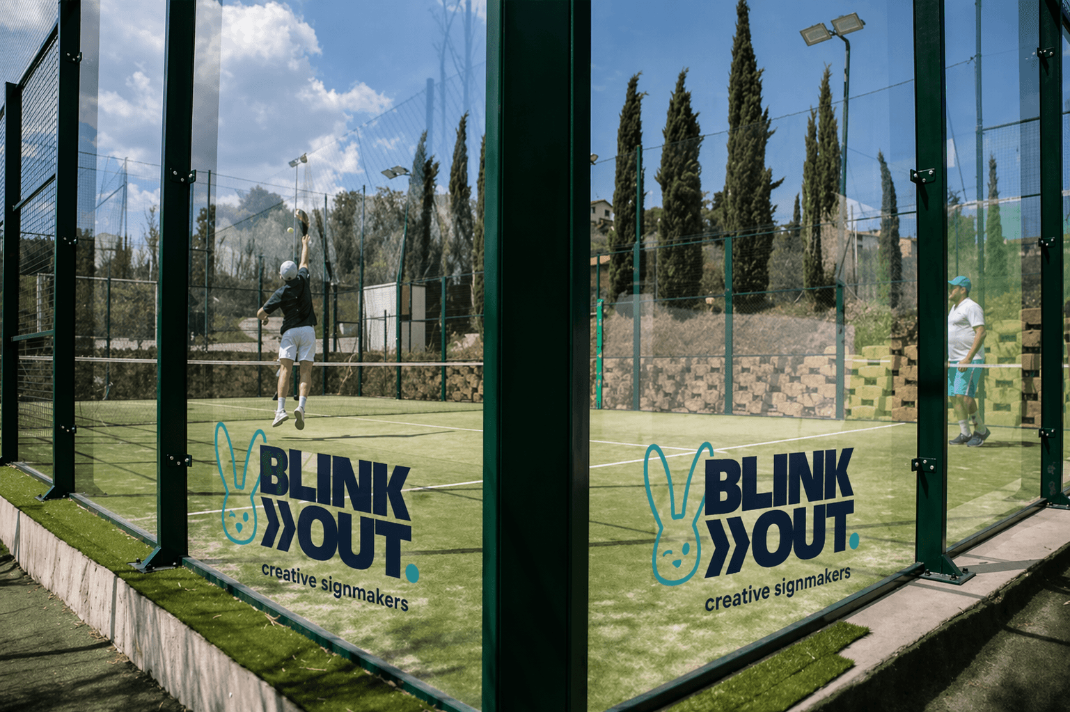 Glaswand van een padelbaan met Blink Out creative signmakers-logo in turquoise en donkerblauw, aangebracht als transparante raamsticker op sportbeglazing.