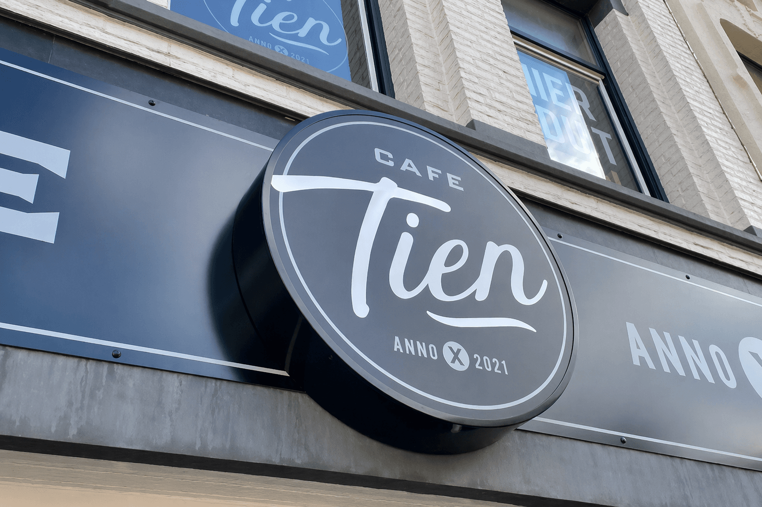 Rond donkerblauw gevelbord van Café Tien met witte belettering en logo, gemonteerd als uithangbord aan de voorgevel van een horecapand.