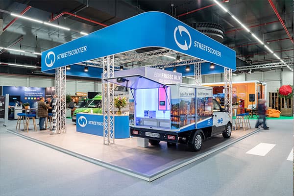 Beursstand van StreetScooter met grote hangende signconstructie en elektrische bakwagen met full-color voertuigbelettering, gepresenteerd in een indoor evenementenhal.