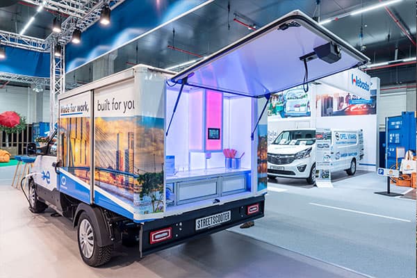 Elektrische StreetScooter bakwagen op een beursstand met opengeklapte laadbak als display, verlichte binnenzijde en full-color voertuigbelettering met stadsbeeld op het zijpaneel.