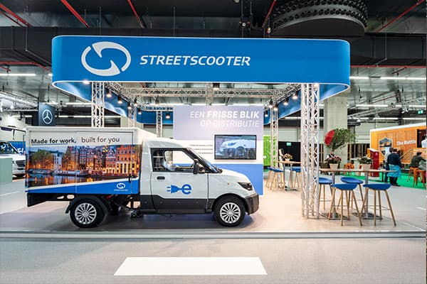 Beursstand van StreetScooter met ronde hangende lichtbak, elektrische bestelwagen met full-color voertuigbelettering en promotionele achterpanelen in een indoor evenementenhal.