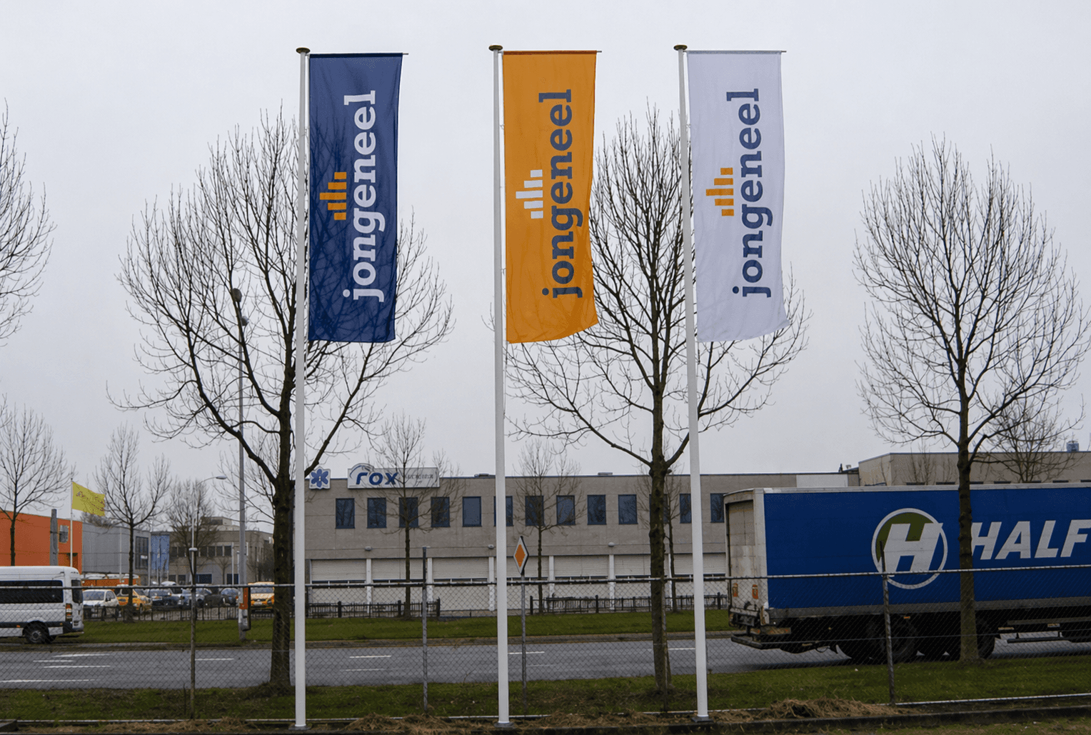 Drie verticale baniervlaggen met Jongeneel-logo in blauw, oranje en wit aan vlaggenmasten langs een bedrijventerrein.