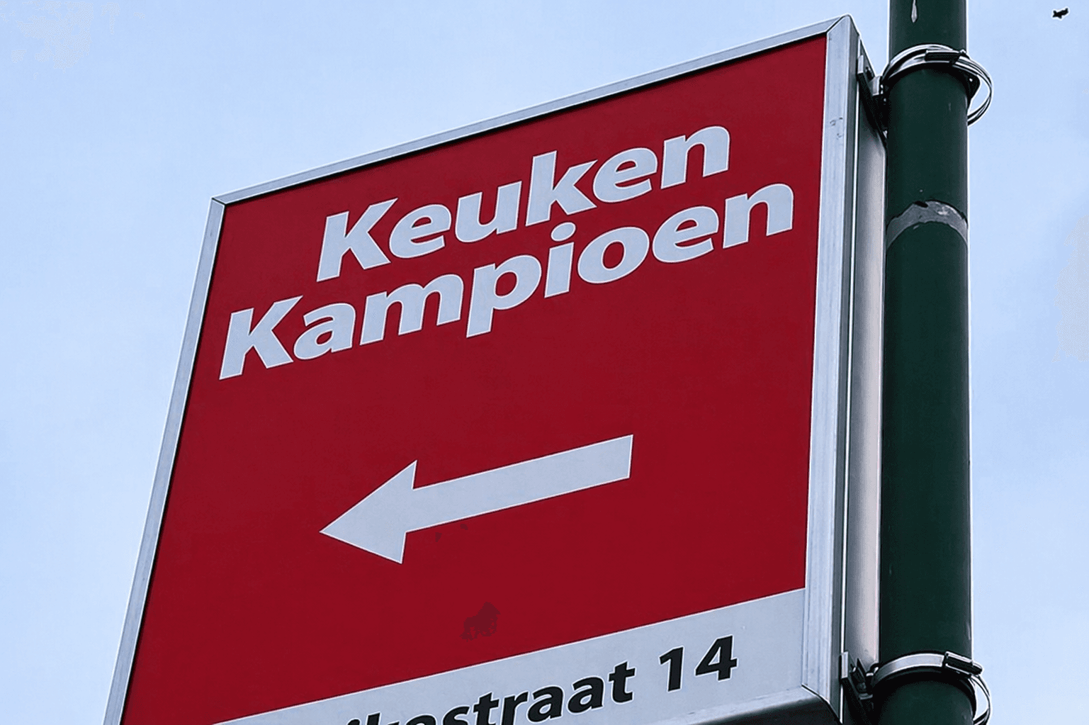 Rood wayfinding-bord van Keuken Kampioen met witte belettering en pijl naar links, gemonteerd aan een groene paal.