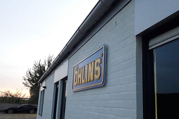 Witte bakstenen gevel met rechthoekig gevelbord van Öhlins in goud en blauw, gemonteerd naast de entree van een bedrijfspand.