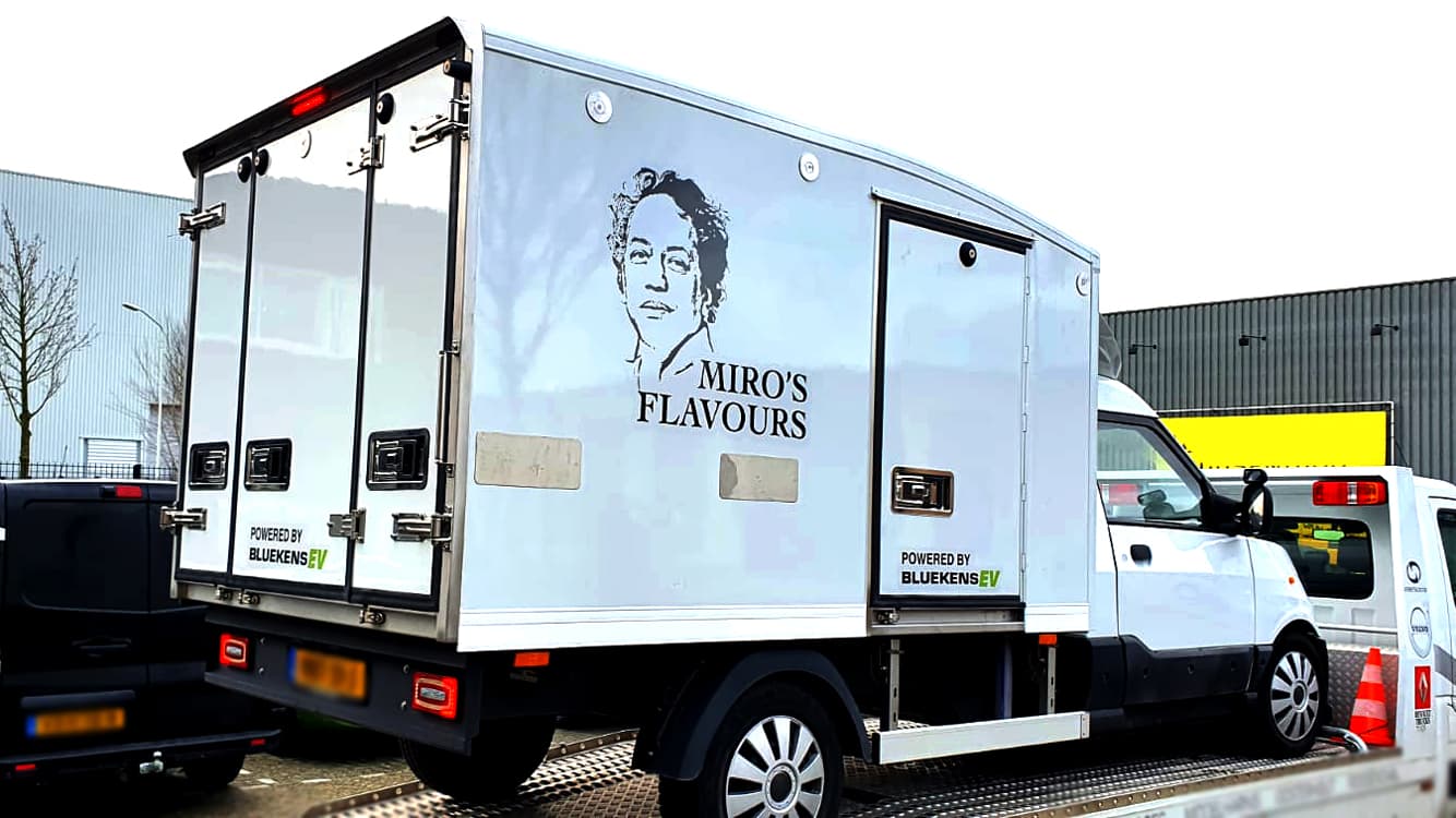 Witte bakwagen met voertuigbelettering voor Miro’s Flavours, voorzien van zwarte merknaam, portretillustratie en sponsorvermelding op de zij- en achterzijde van de laadbak.