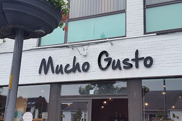 Witte bakstenen winkelgevel met zwarte sierlijke gevelletters "Mucho Gusto" boven de glazen entree van een horecapand.