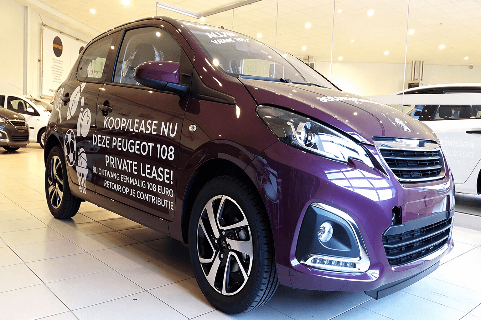 Paarse Peugeot 108 met witte voertuigbelettering en promotionele autobelettering op de zijkant, gefotografeerd in een showroom met duidelijke reclameletters en vinylstickers.