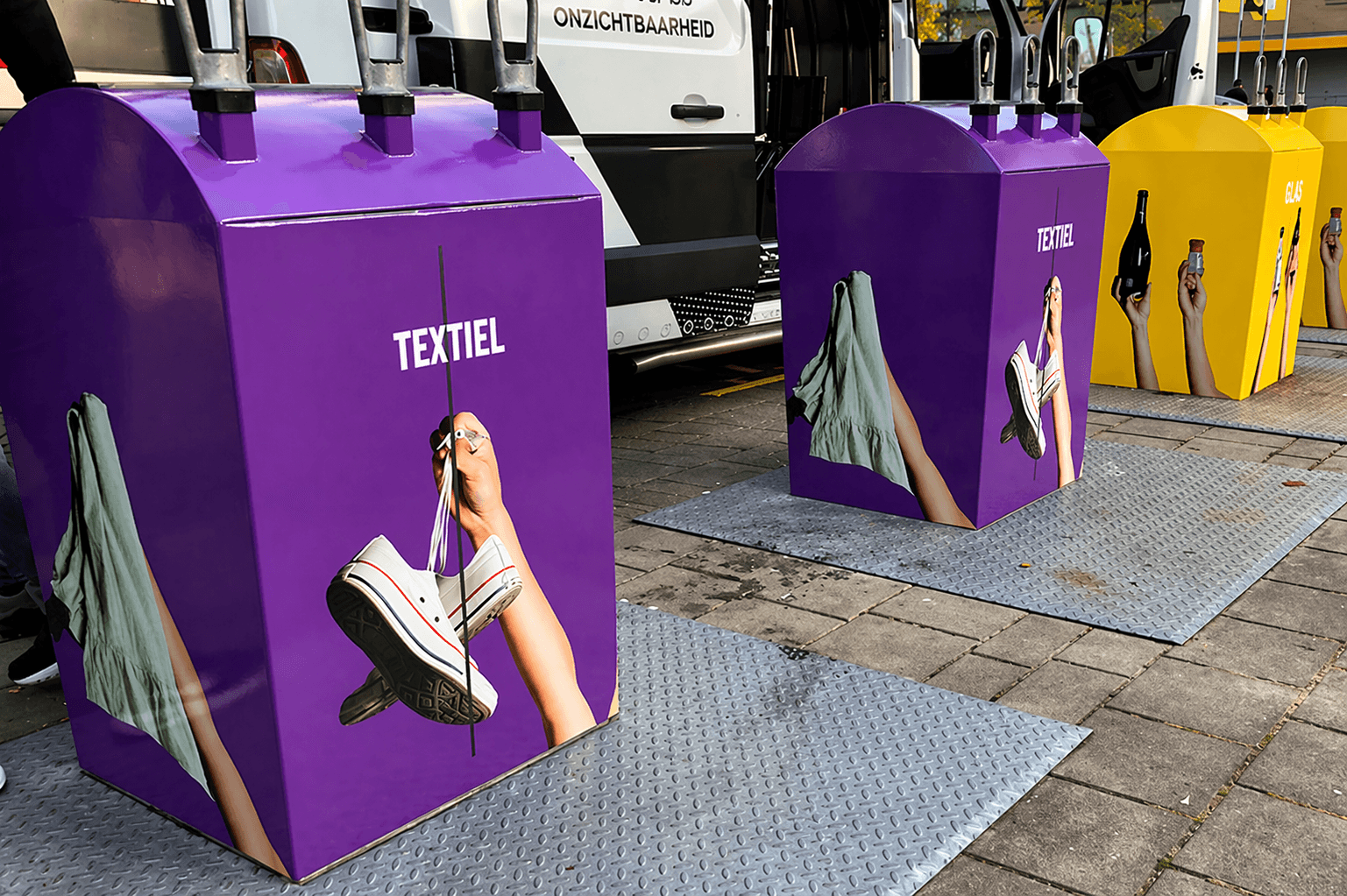 Full colour containerbelettering op paarse textielcontainers en gele glascontainers tijdens plaatsing in een openbare buitenruimte.