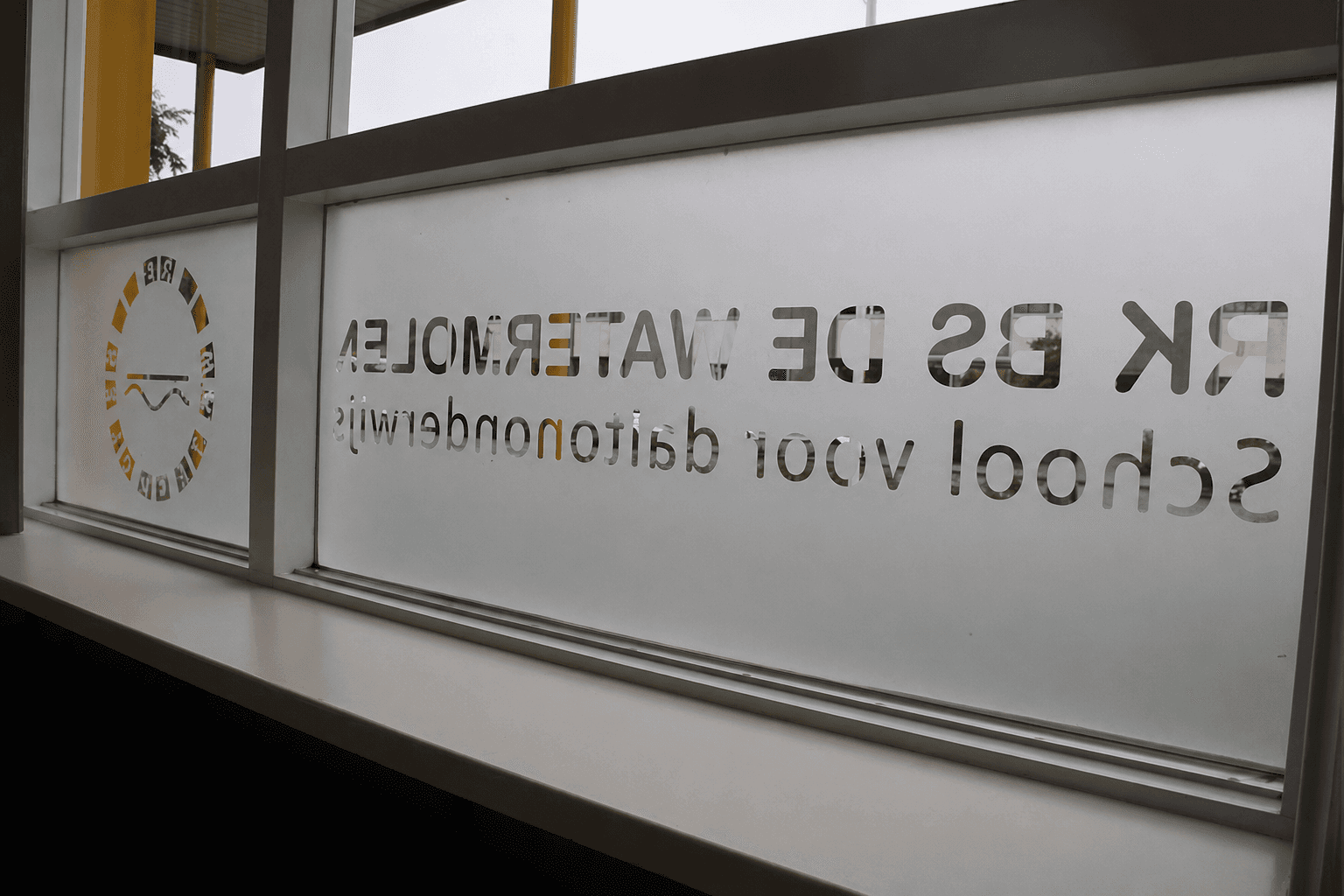 Glasfolie met uitgespaarde schoolnaam en rond logo op een raam, toegepast als raamdecoratie en privacyfolie.