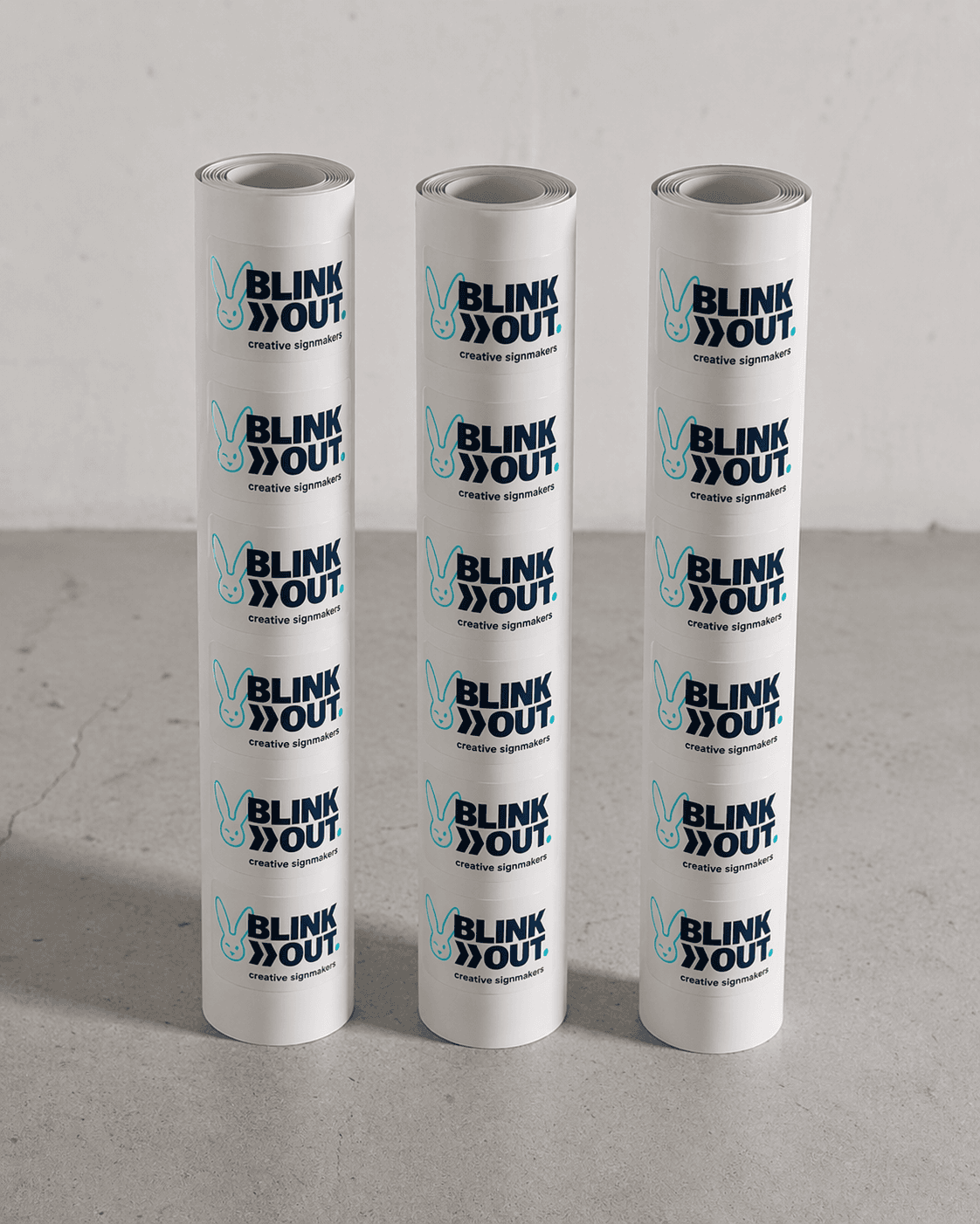 Drie rollen witte etiketten met herhaald BLINK OUT creative signmakers-logo, gepresenteerd als bedrukte stickers op een neutrale ondergrond.