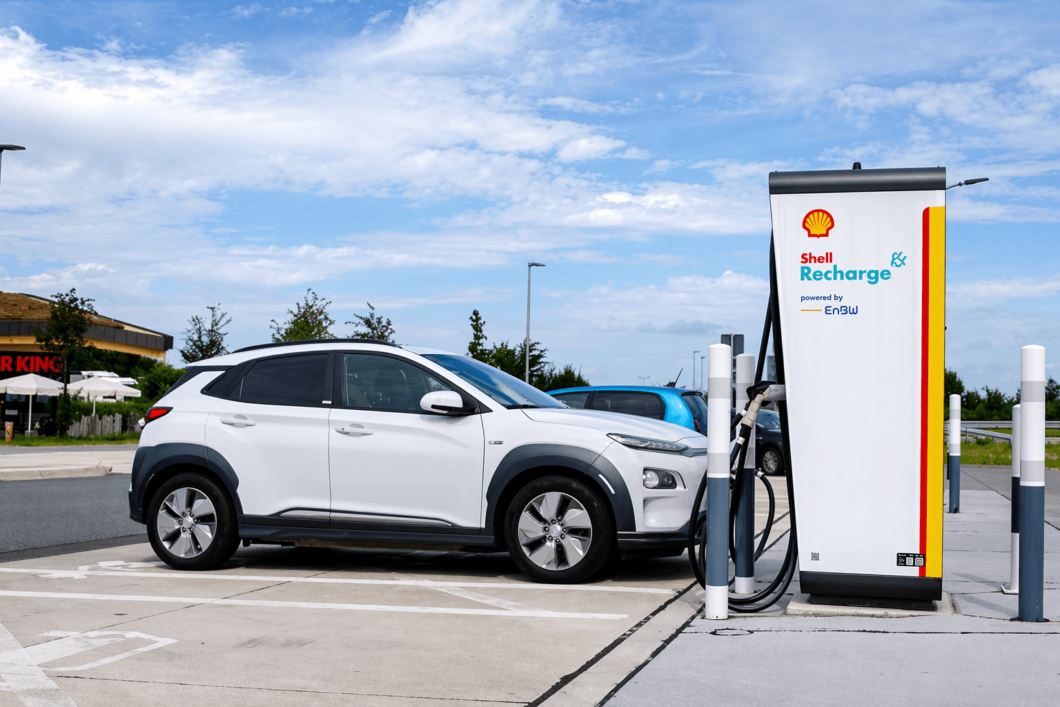 Shell Recharge laadpaal met branding bij een snellaadstation waar een witte elektrische auto wordt opgeladen.