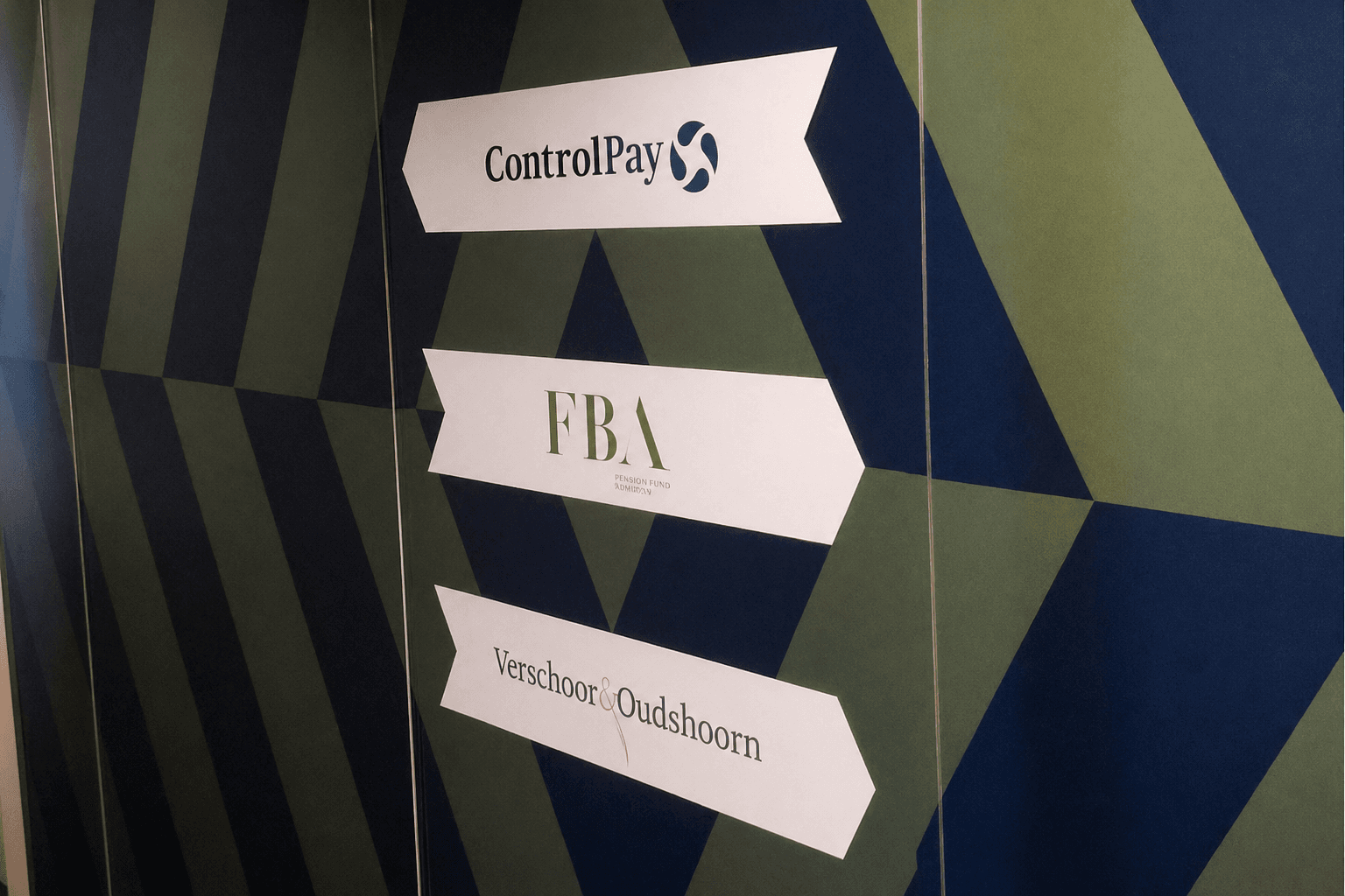 Wayfinding-wand met geometrische wandbekleding en witte richtingsborden voor ControlPay, FBA en Verschoor & Oudshoorn als kantoorbewegwijzering.
