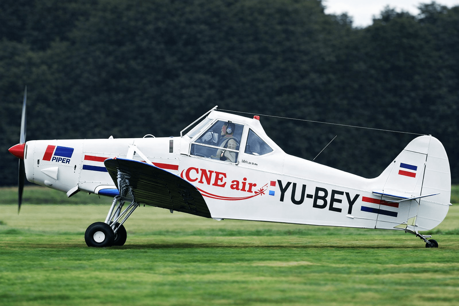 Wit sportvliegtuig met Piper-logo, CNE air-belettering en grote registratiemarkering YU-BEY op de romp, gefotografeerd op een grasveld met bosrand op de achtergrond.