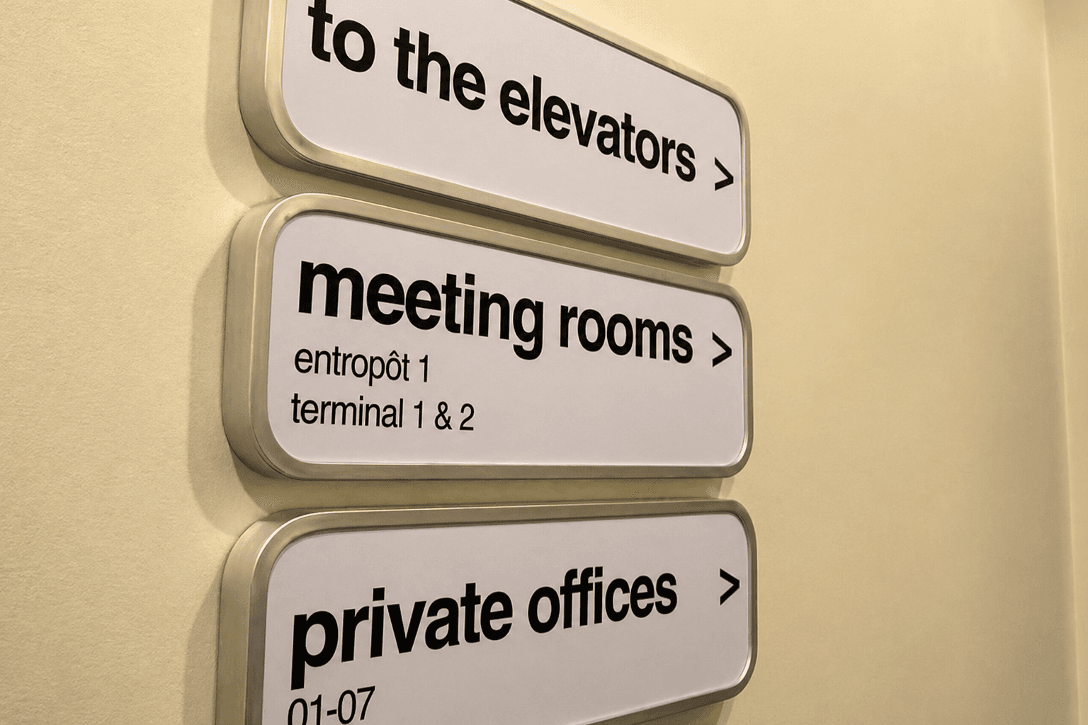 Wandgemonteerde wayfinding-borden met aluminium frame en zwarte tekst voor liften, vergaderruimtes en kantoren op een lichte binnenmuur.