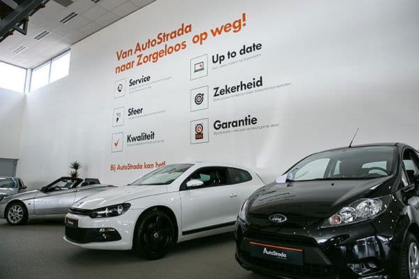 Interieur van een autoshowroom met grote wandbelettering en informatieve muurgrafiek van AutoStrada, met kernwaarden in zwarte en oranje tekst boven meerdere showroomauto’s.