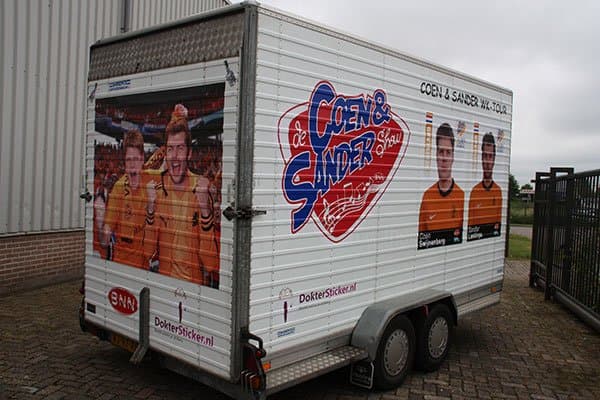 Gesloten tandemasser aanhangwagen met full-colour bestickering, grote fotoprints van twee mannen in oranje shirts en het logo van de Coen & Sander Show op zij- en achterzijde.