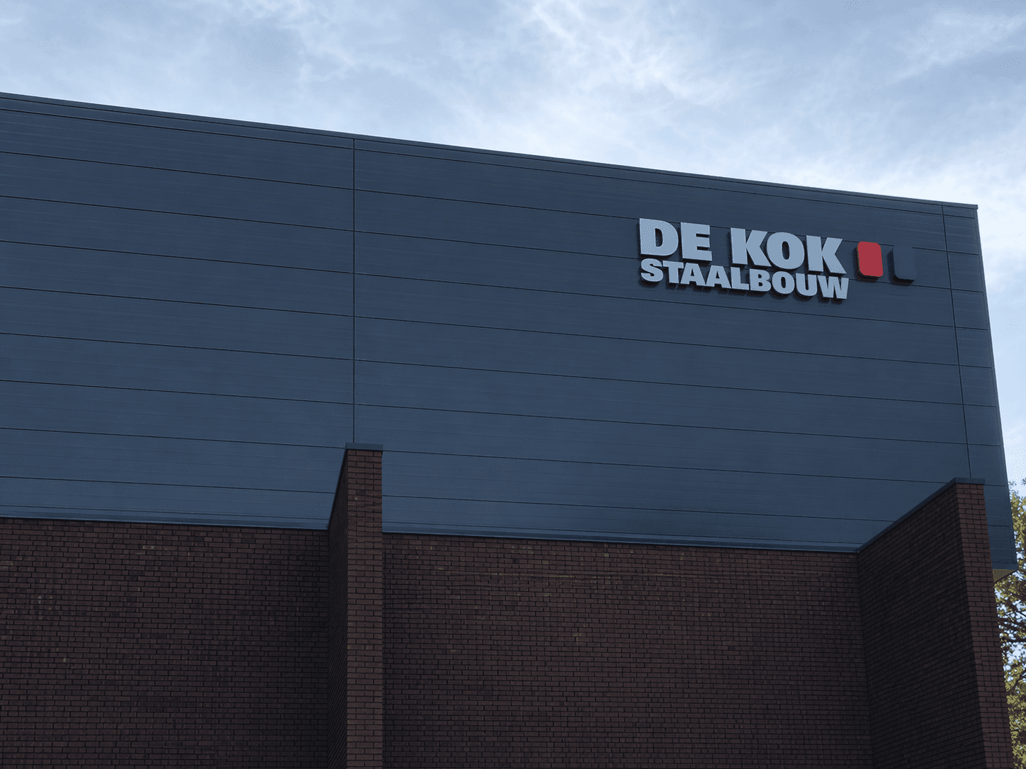 Moderne bedrijfshal met gevelreclame van DE KOK STAALBOUW in witte 3D-letters en rood-blauw logo op donkergrijze gevelpanelen boven een bakstenen plint.