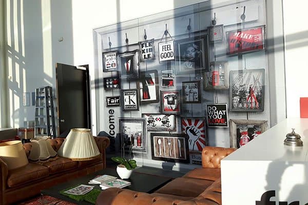 Loungehoek in een modern bedrijfsinterieur met leren banken en een grote full-color glaswandprint in collage-stijl met typografie, posters en framed artwork.