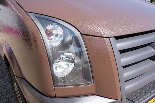 Close-up van de voorzijde van een bestelbus met bruine carwrap, zichtbare folietextuur, koplampdetail en zilverkleurige grille.