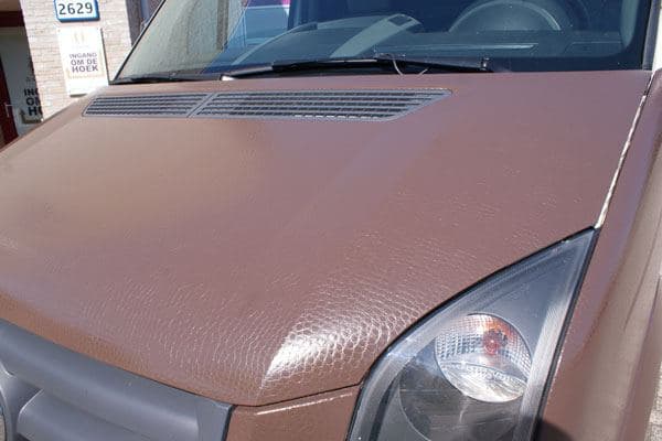 Close-up van de motorkap van een bestelbus met bruine carwrap, zichtbare folietextuur, ventilatierooster en koplampdetail aan de voorzijde.