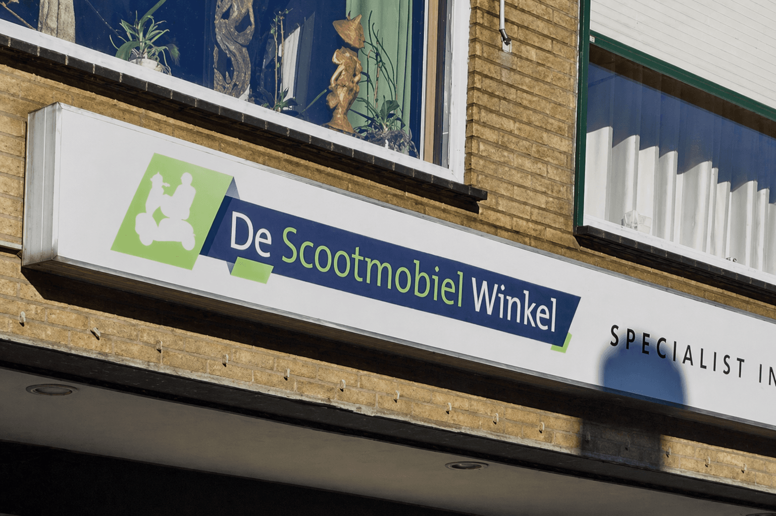 Wit gevelbord met groen-blauw logo en belettering van De Scootmobiel Winkel, gemonteerd op een bakstenen winkelgevel onder de ramen.