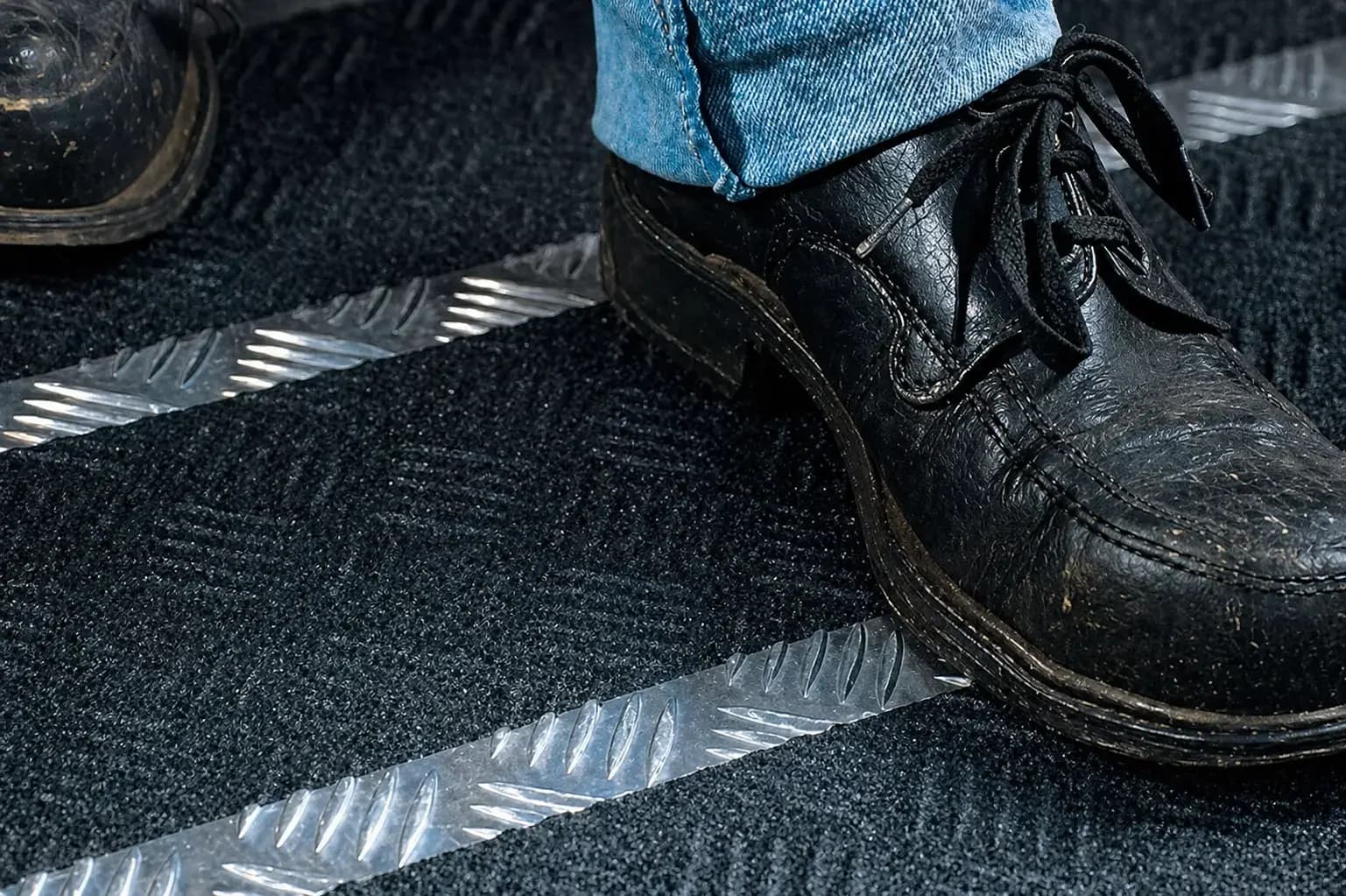 Close-up van zwarte schoenen op een donkere vloermat met zilverkleurige antislipstrips, toegepast als veiligheidsmarkering en slippreventie op een looproute.
