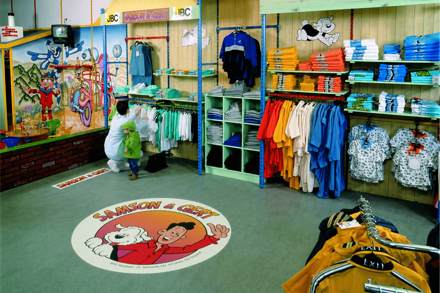 Kinderkledingwinkel met kleurrijke Samson & Gert wanddecoratie, schapbordjes, vloersticker en thematische signing voor een speelse winkelinrichting.