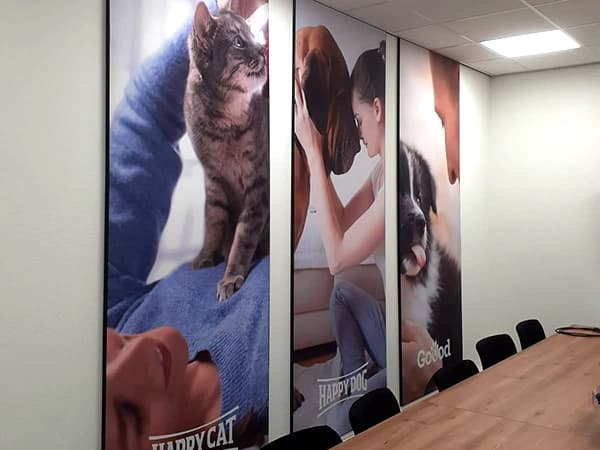 Vergaderruimte met grote wandvisuals van katten, honden en baasjes op verticale textielframes, toegepast als full-colour interieurbelettering.