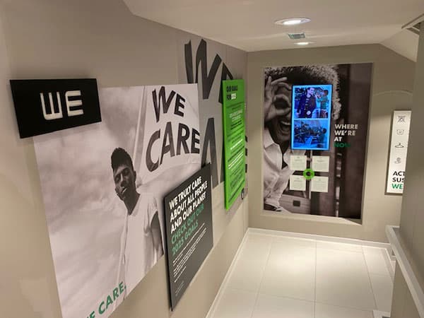 Trappenhuis met grote wandvisuals en interieurbelettering met de slogan "WE CARE", gecombineerd met fotopanelen en informatieve muurcommunicatie in een moderne kantooromgeving.
