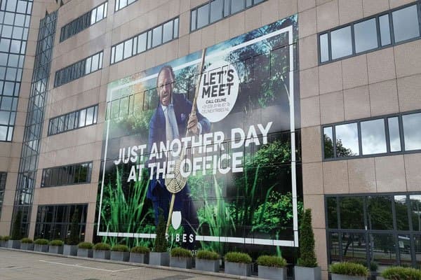Kantoorgebouw met een gigantisch full-color geveldoek over meerdere verdiepingen, voorzien van een campagnebeeld en grote witte typografie “Just another day at the office”.