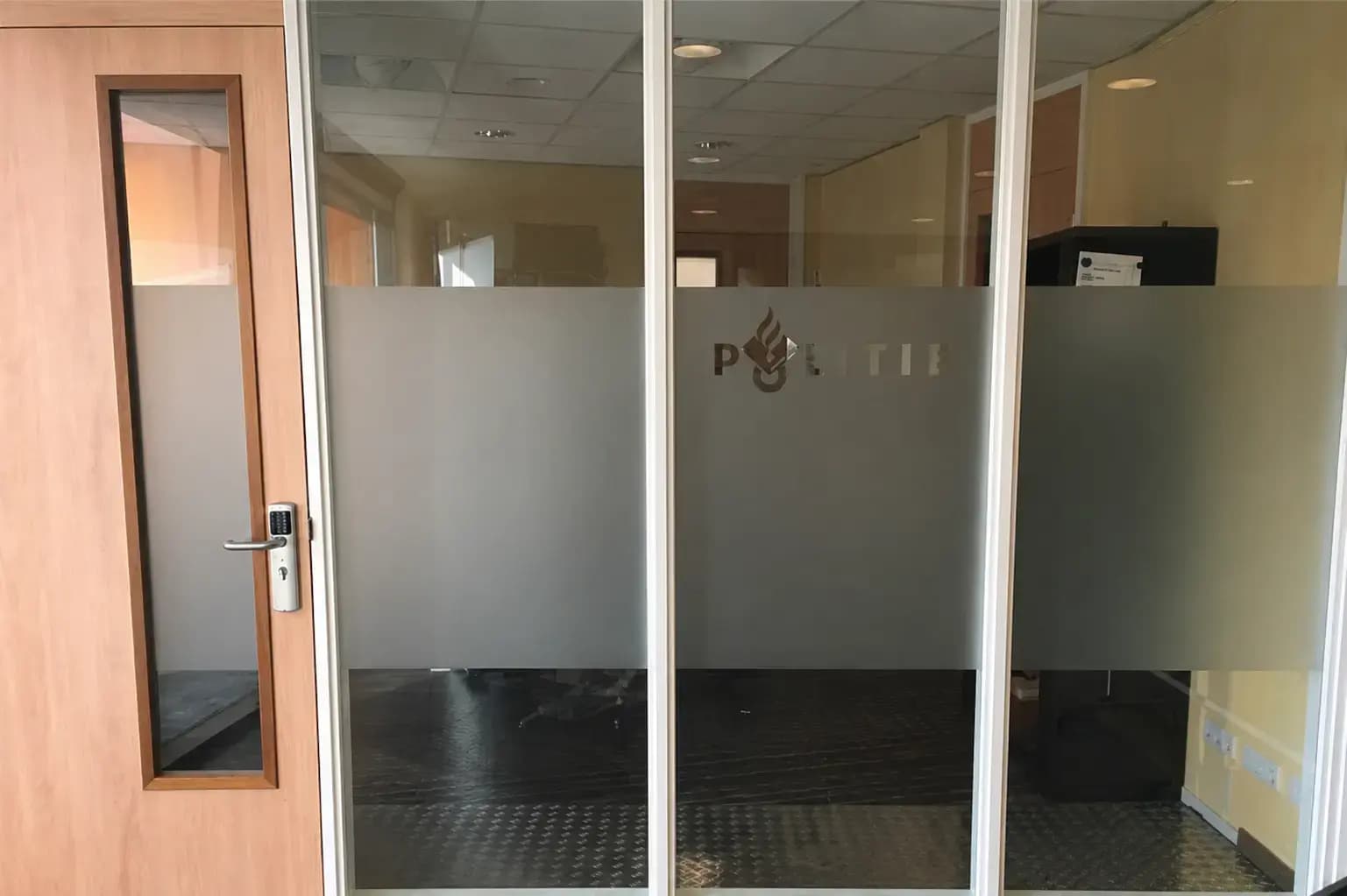 Glaswand met brede matte privacyfolie en uitgesneden Politie-logo, toegepast als functionele kantoorbelettering en afscherming op een binnenpui.
