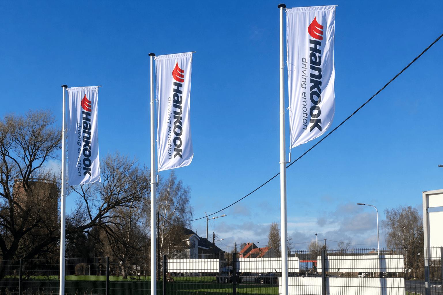 Drie witte mastvlaggen met Hankook-logo en verticale belettering, geplaatst op hoge vlaggenmasten op een bedrijventerrein onder een heldere blauwe lucht.