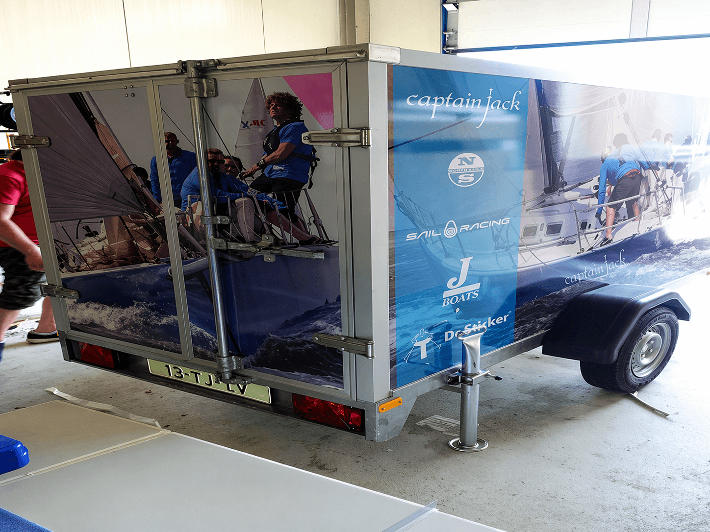 Volledig bestickerde gesloten aanhangwagen met full-colour voertuigbelettering, zeilbootfotoprints en sponsorlogo’s, gefotografeerd in een werkplaats.