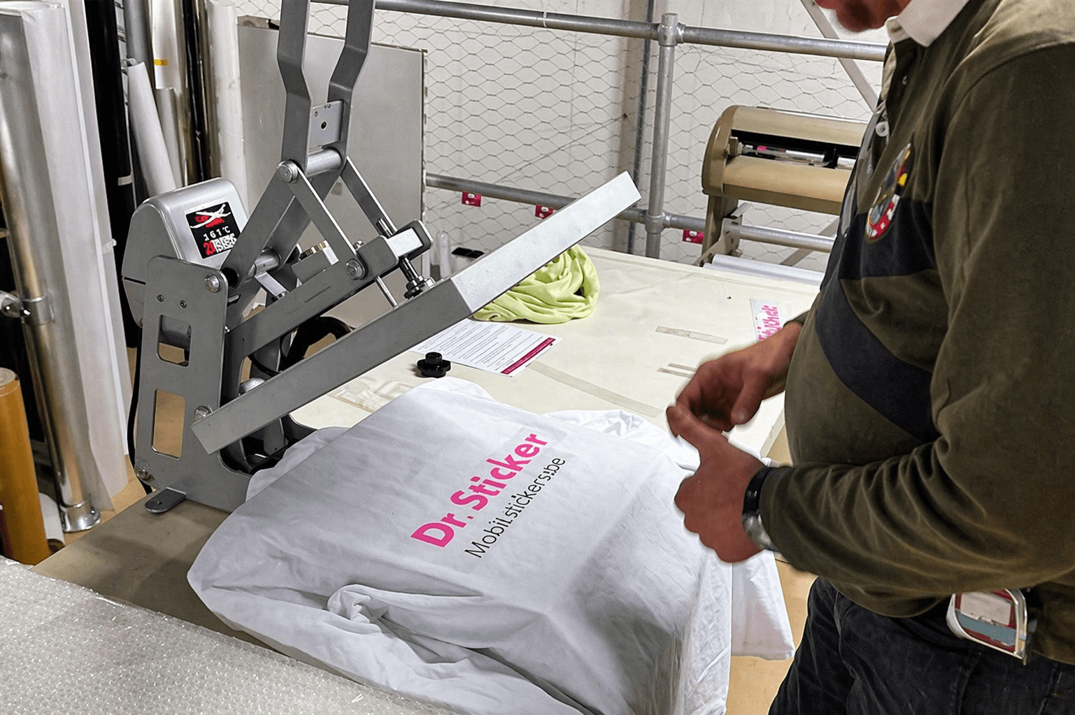 Werkplaatsbeeld van textielbedrukking met hittepers, waarbij een witte T-shirt met roze-zwarte Dr. Sticker-belettering wordt gepositioneerd voor transferdruk.