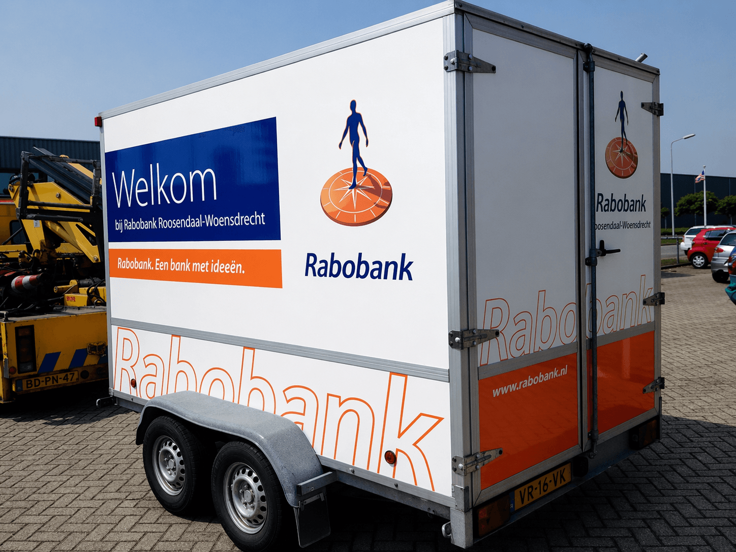 Dubbelassige gesloten aanhanger met full colour voertuigbelettering van Rabobank, inclusief welkomsttekst, logo en oranje-blauwe huisstijl op de zijkant en achterdeuren.