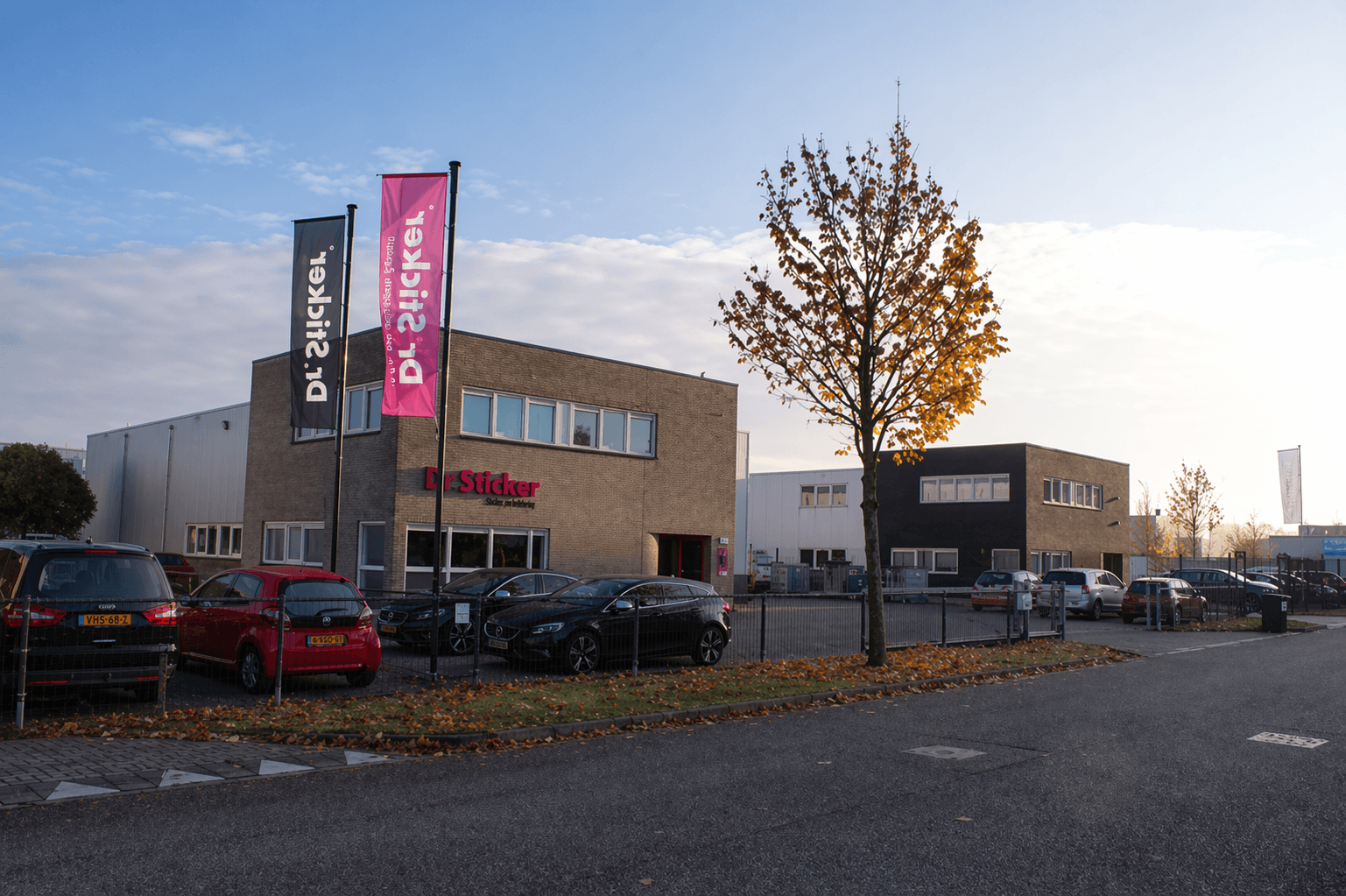 Bedrijfspand op een bedrijventerrein met gevelreclame van Dr. Sticker en twee hoge mastvlaggen in donkerblauw en magenta, gefotografeerd langs een straat met geparkeerde auto’s.