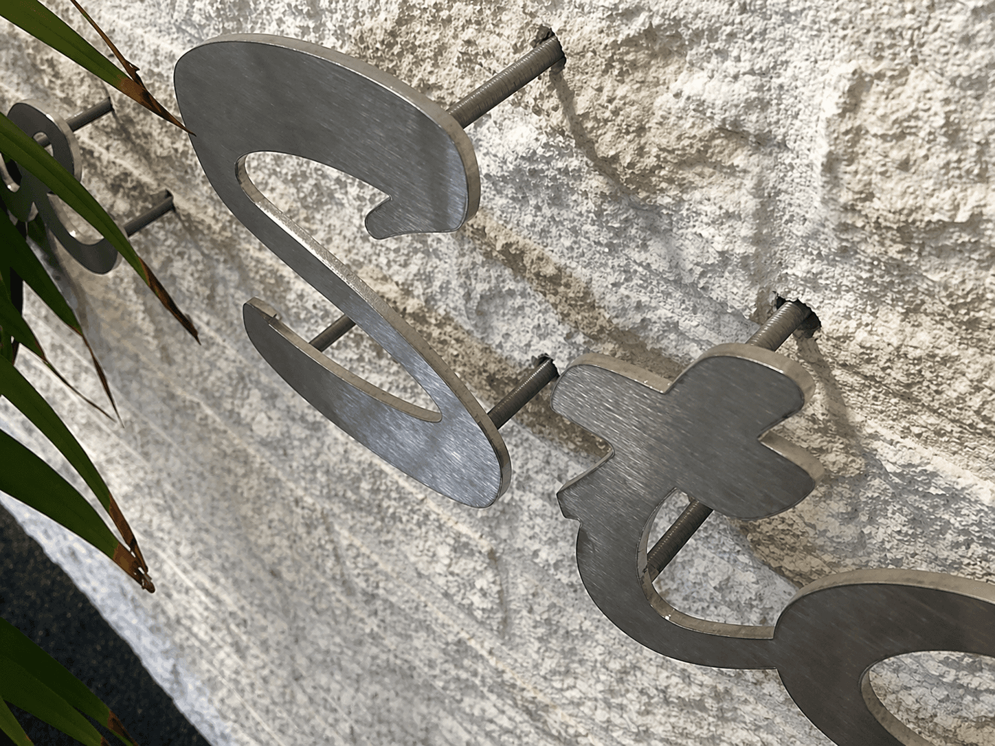 Close-up van geborstelde metalen gevelletters op afstandhouders, gemonteerd op een ruwe natuurstenen wand als luxe 3D-belettering.