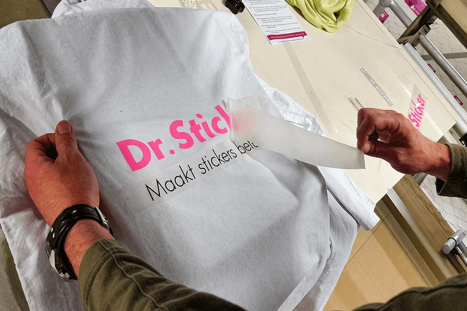 Handen brengen roze-zwarte textielbelettering met de tekst Dr. Stick aan op een wit T-shirt door transferfolie te verwijderen tijdens het bedrukken van bedrijfskleding.