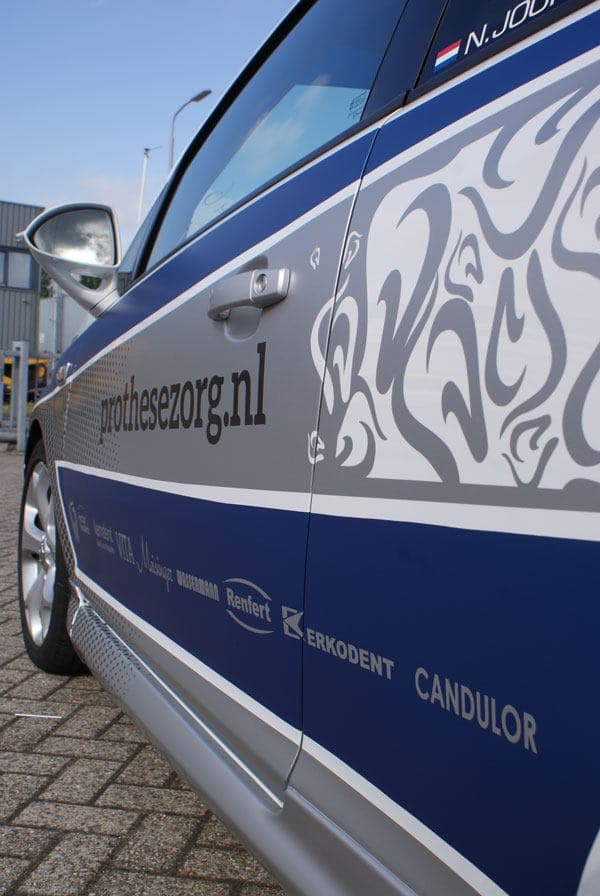 Close-up van blauwe en grijze voertuigbelettering op een witte personenauto, met prothesezorg.nl, decoratief patroon en sponsorlogo’s op het portier.